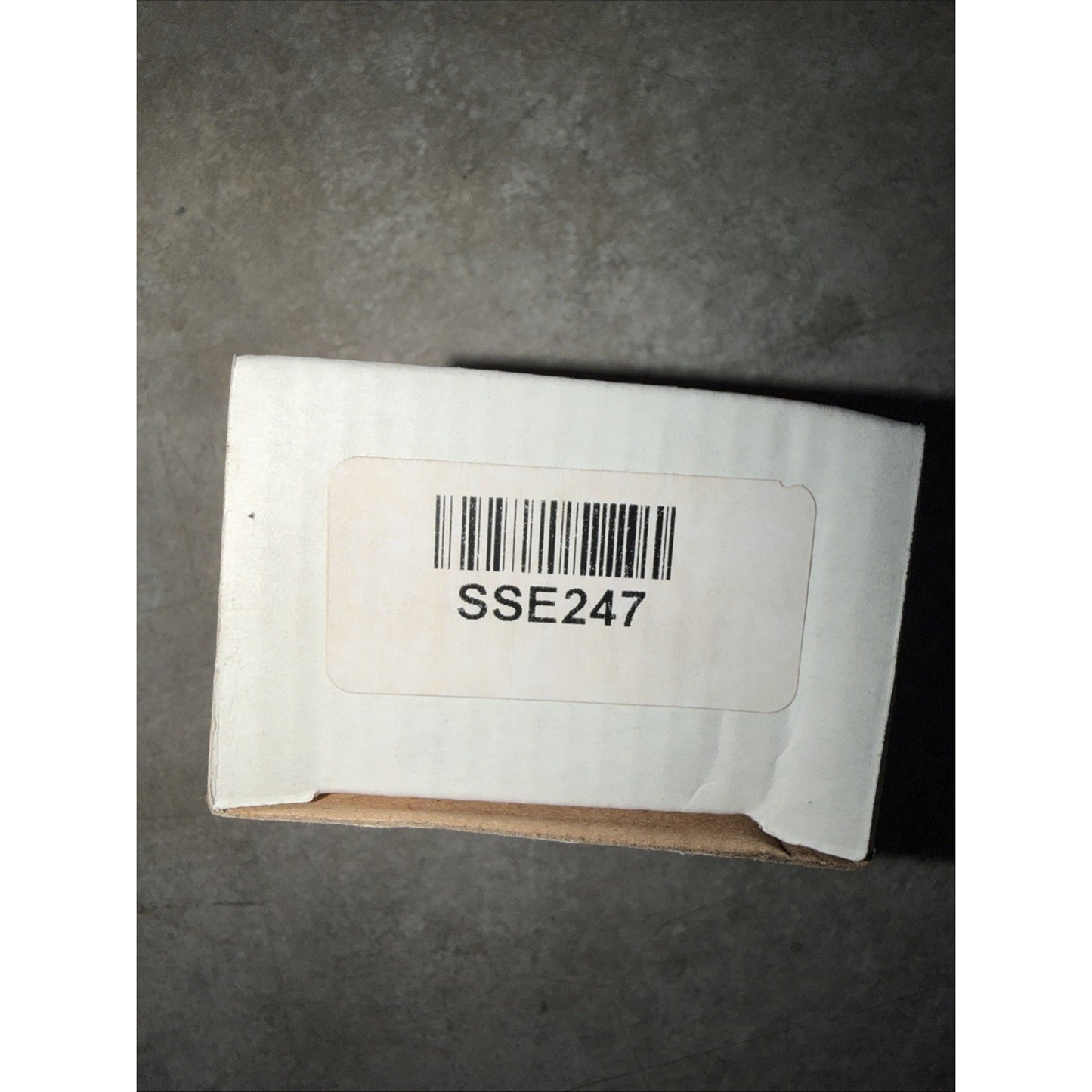 Small plain white cardboard box with a sticker label showing a barcode and the code "SSE247" on a gray surface background.