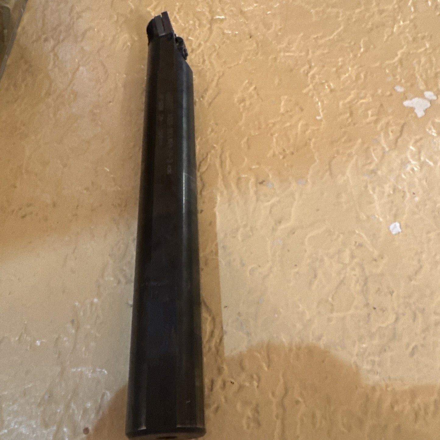 Dark cylindrical metal tool with a small cutting tip and clamp mechanism on one end, positioned vertically against a textured beige wall background.