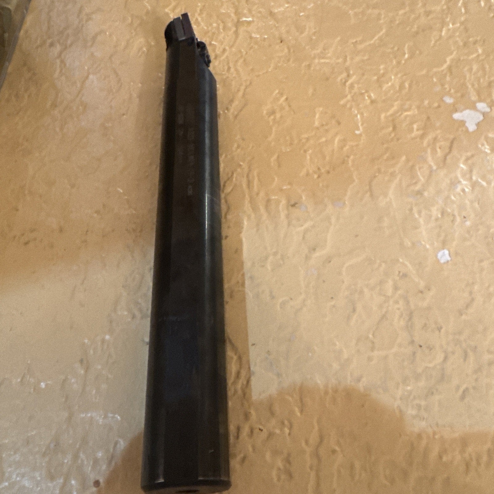 Dark cylindrical metal tool with a small cutting tip and clamp mechanism on one end, positioned vertically against a textured beige wall background.