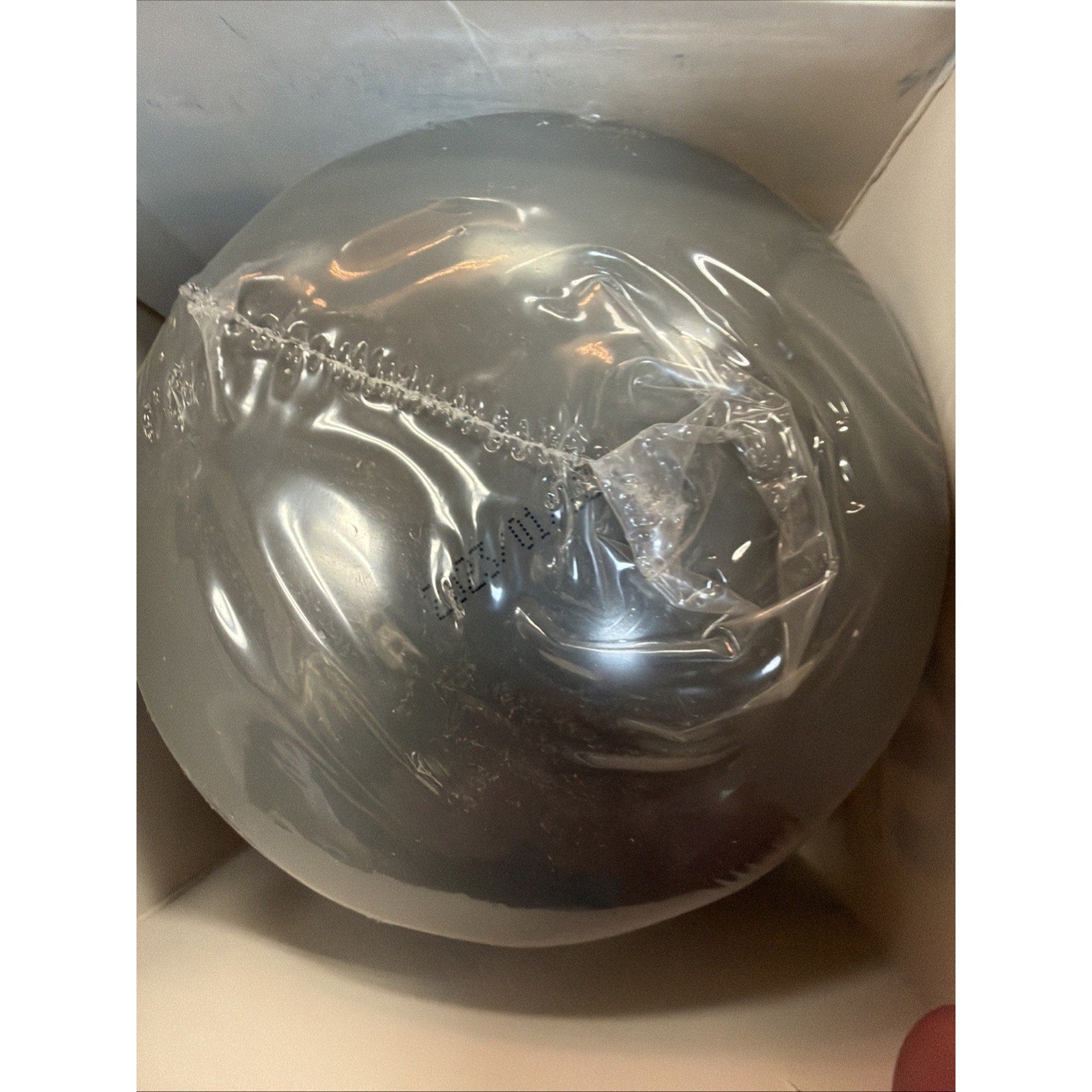 Silver-colored spherical object tightly wrapped in clear plastic inside a white cardboard box with visible texture and reflections on the plastic surface.