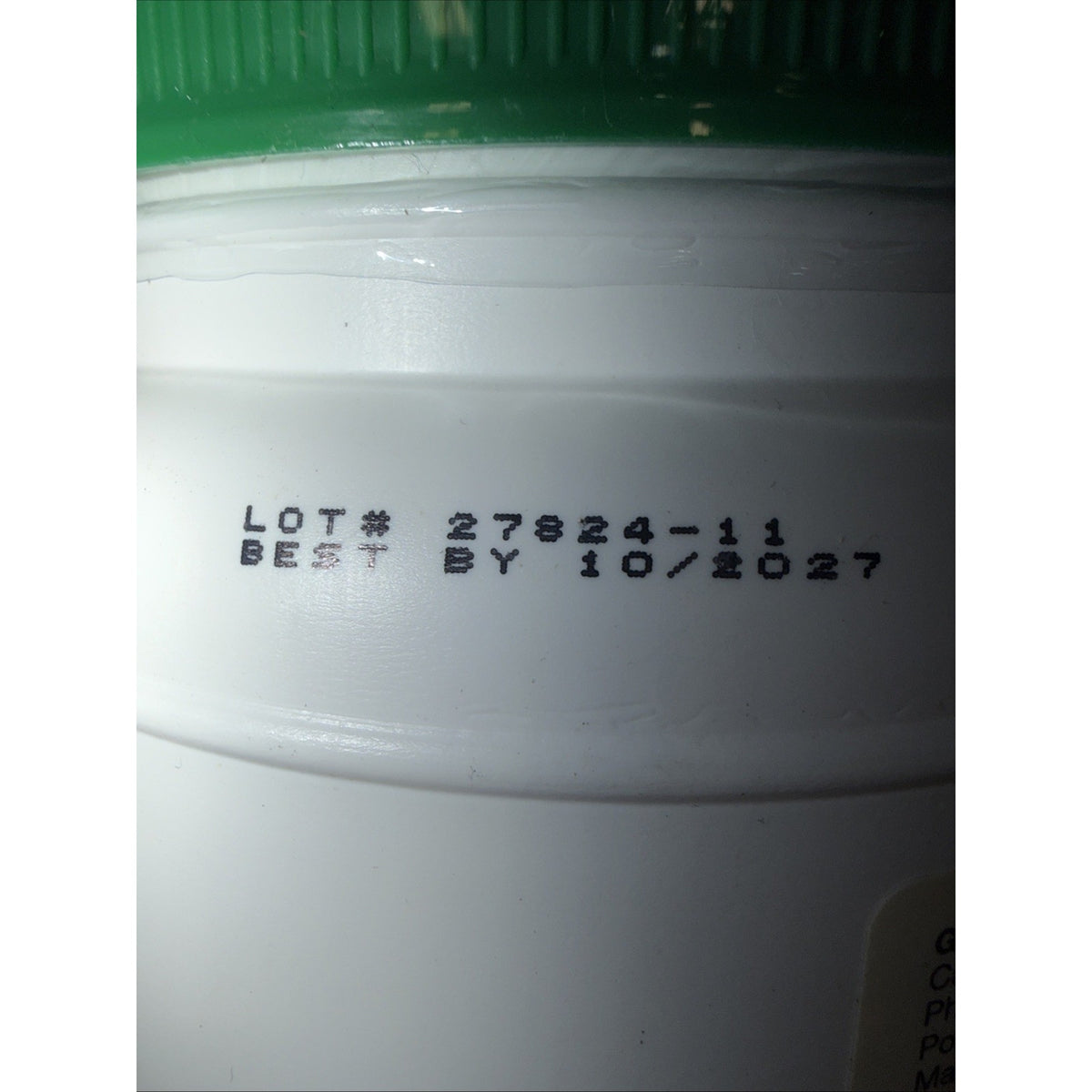Close-up of a white plastic container with a green lid showing a black printed lot number 27824-11 and a best-by date of 10/2027 on the side.