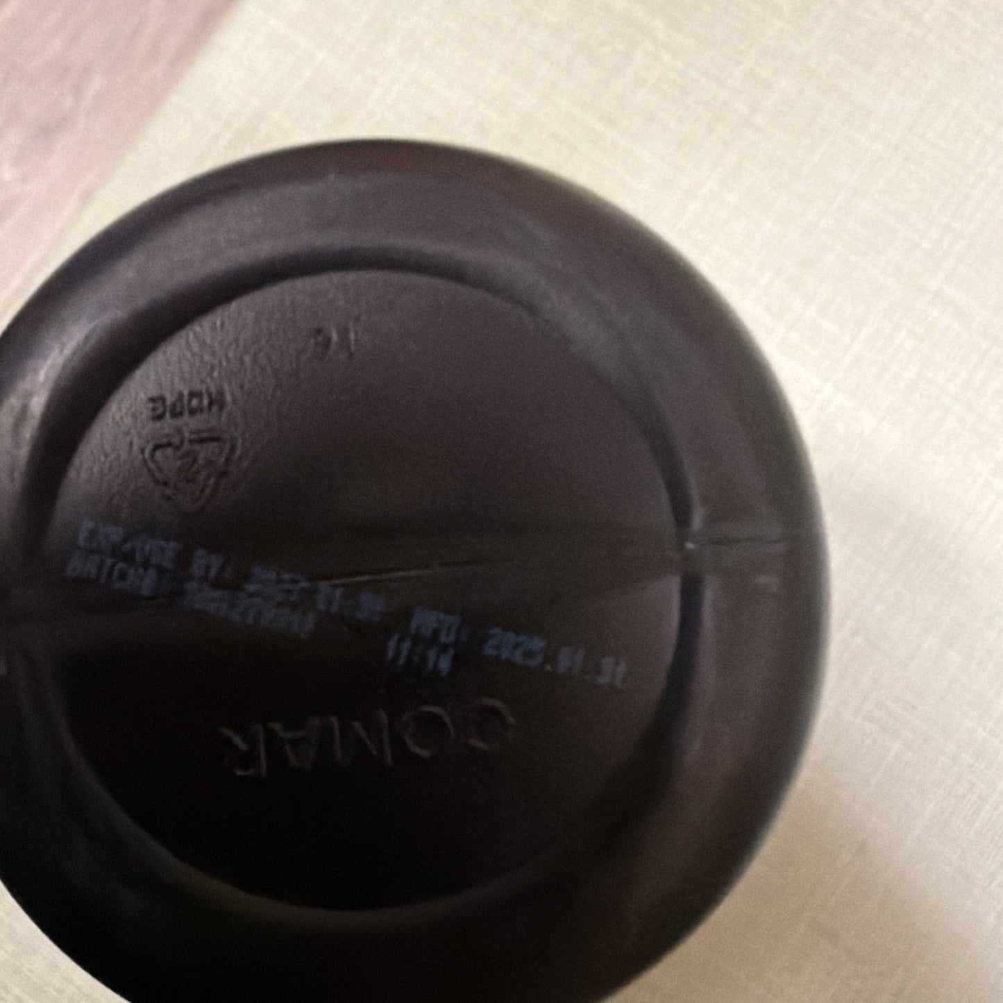 Bottom view of a black plastic container featuring embossed text "COMAR," recycling triangle symbol with code, and faint blue printed manufacturing and expiration date details.
