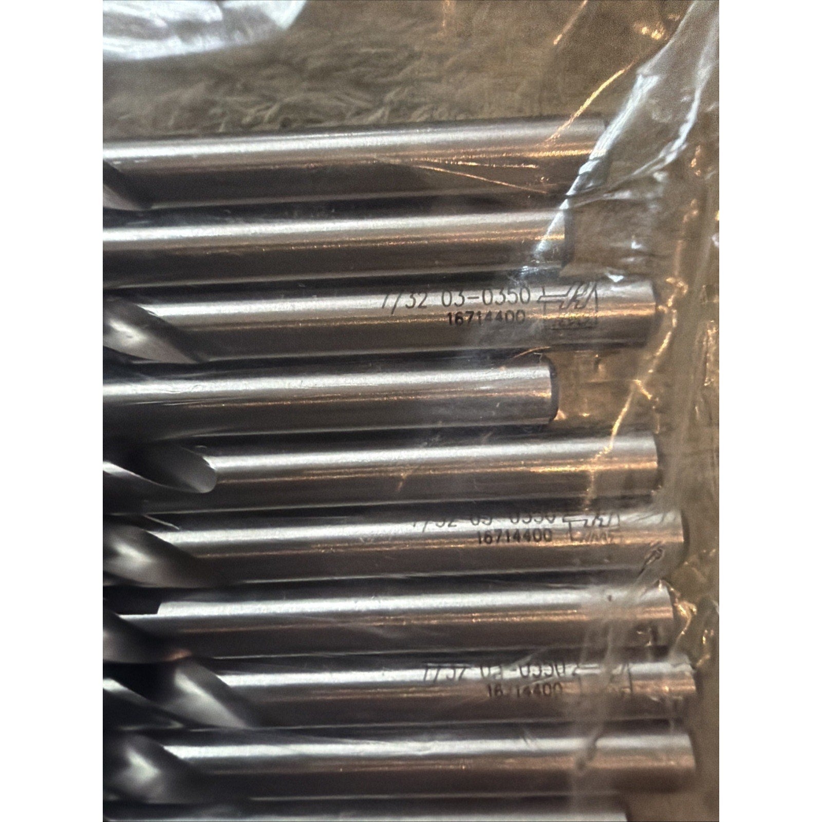 Close-up view of seven silver metal drill bits with engraved size and serial numbers, partially wrapped in clear plastic on a dark textured surface.