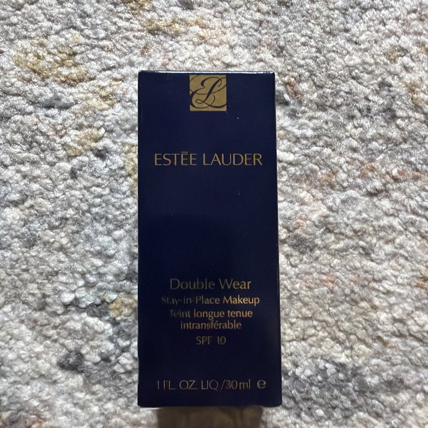 Dark navy blue box of Estée Lauder Double Wear Stay-in-Place Makeup with gold lettering, indicating SPF 10 and 1 fl oz (30 ml) size, placed on a textured carpet surface.