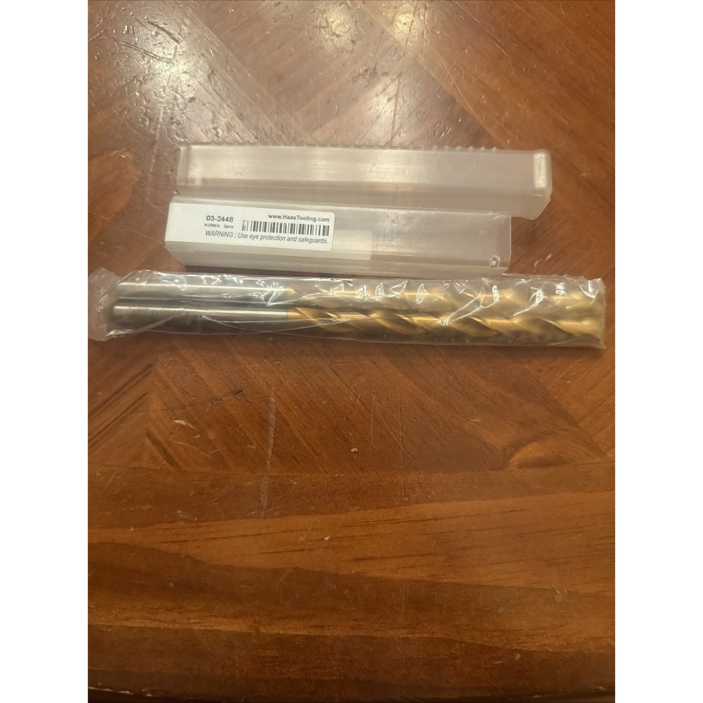 Long metal drill bit with a smooth shank and spiral flutes, sealed in clear plastic packaging, displayed on a wooden surface alongside a rectangular translucent plastic case with a barcode label.