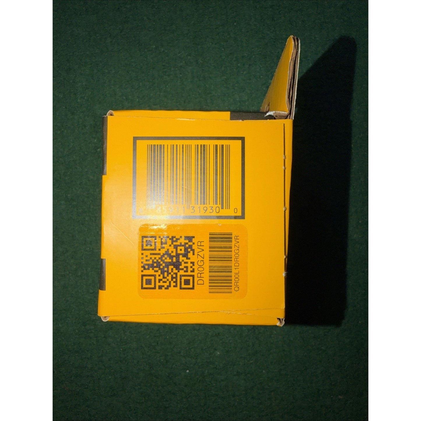 Yellow product box with a barcode and a QR code printed on one side, set against a dark green background with the top flap partially open.