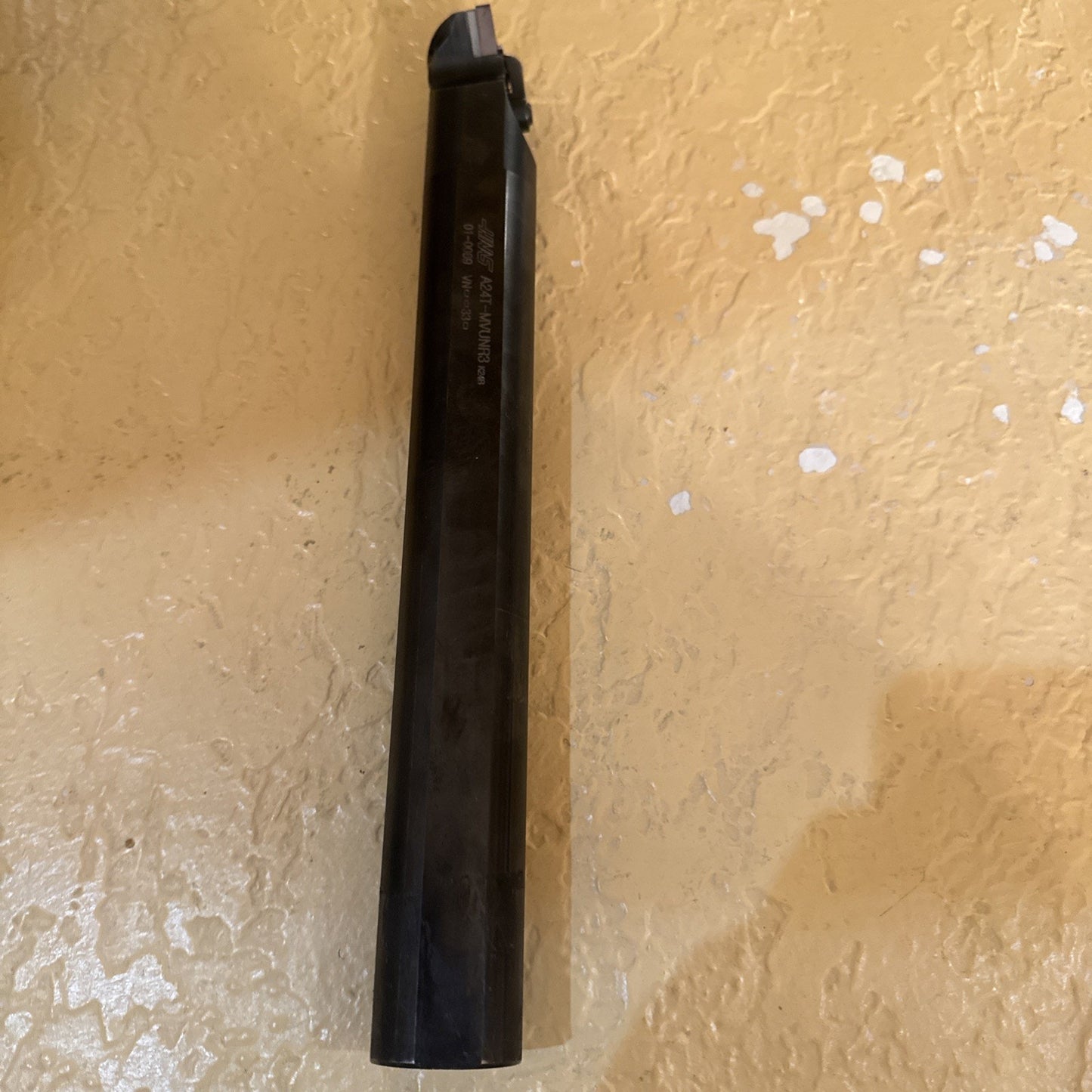 Long black metal cutting tool with a square-shaped tip resting on a textured beige surface, showing faint engraved text along the shaft.