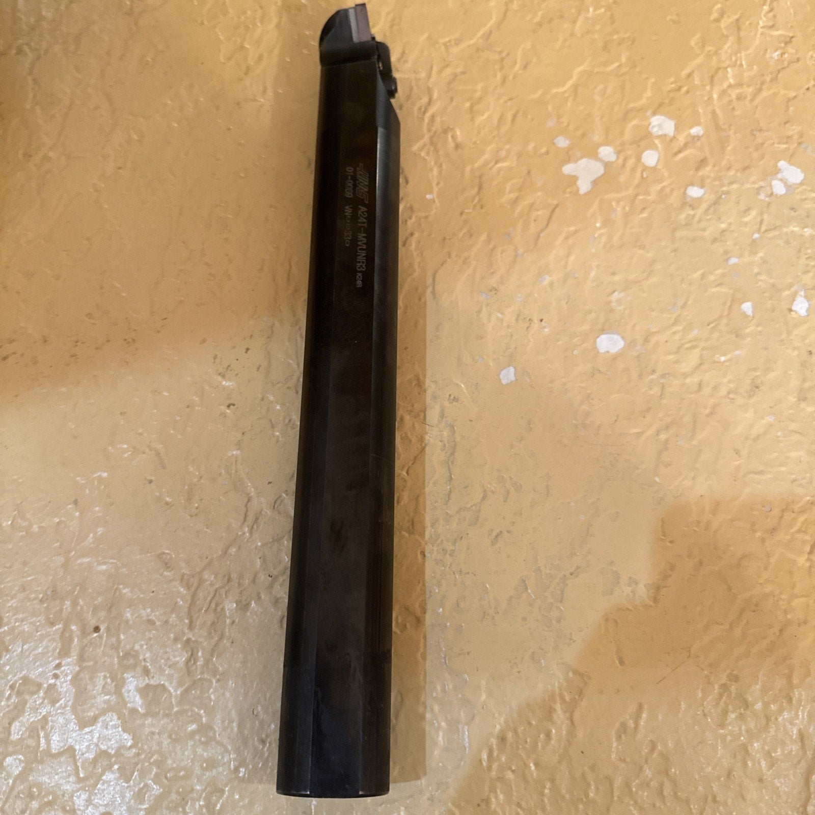 Long black metal cutting tool with a square-shaped tip resting on a textured beige surface, showing faint engraved text along the shaft.