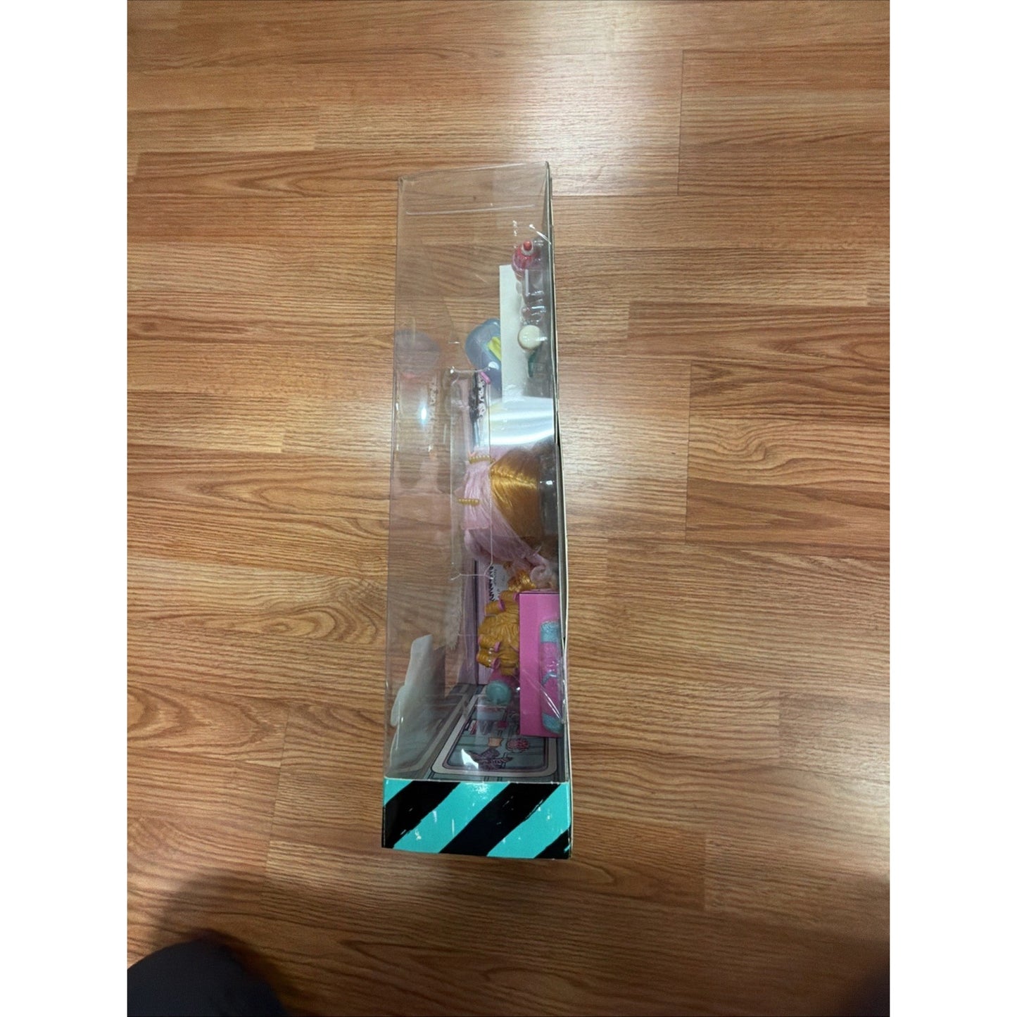 Side view of a clear plastic toy package displaying colorful doll accessories and figures, set against a hardwood floor background.