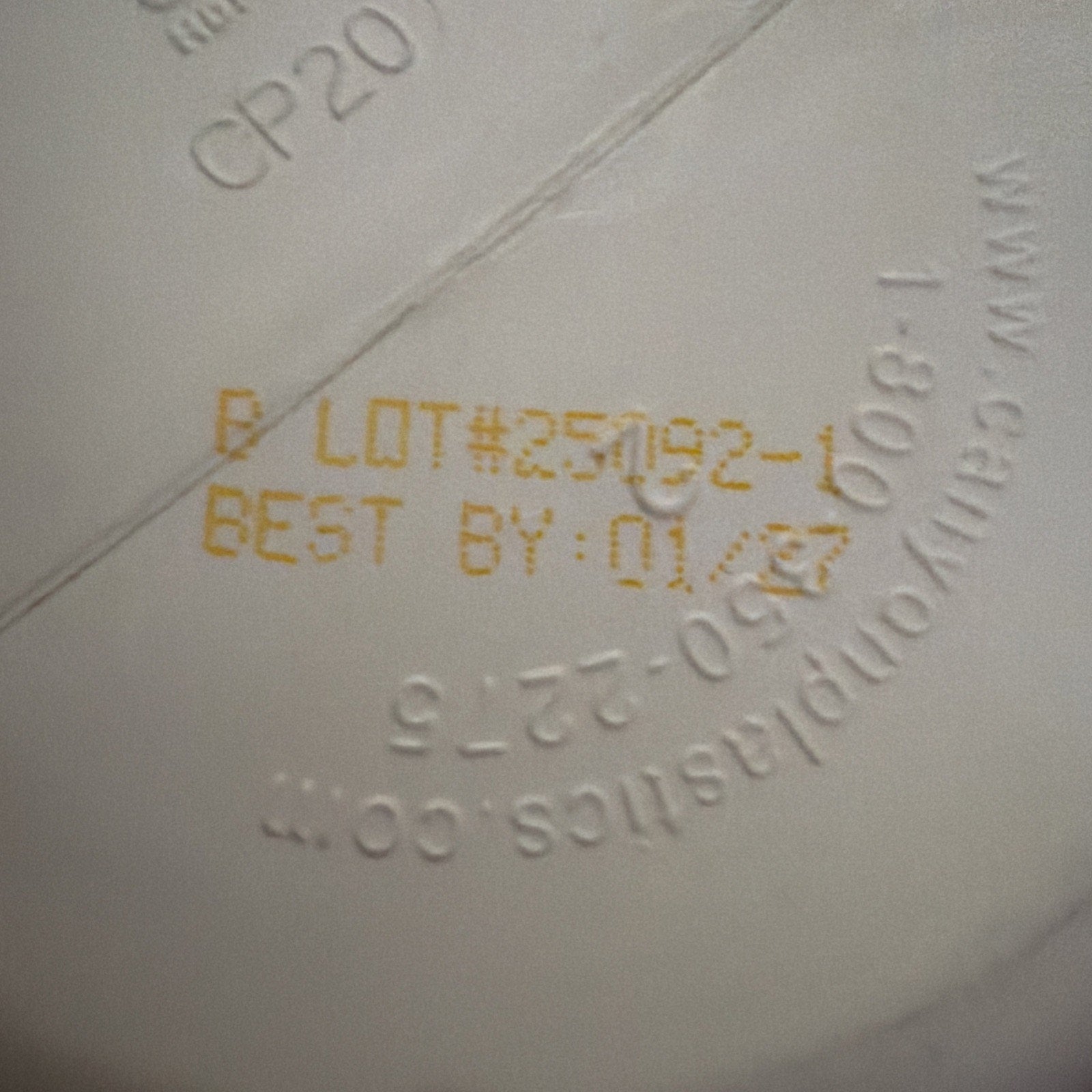 Close-up of a white container bottom showing an embossed website URL and printed yellow batch number "25092-1" with best-by date "01/27" on a smooth surface.