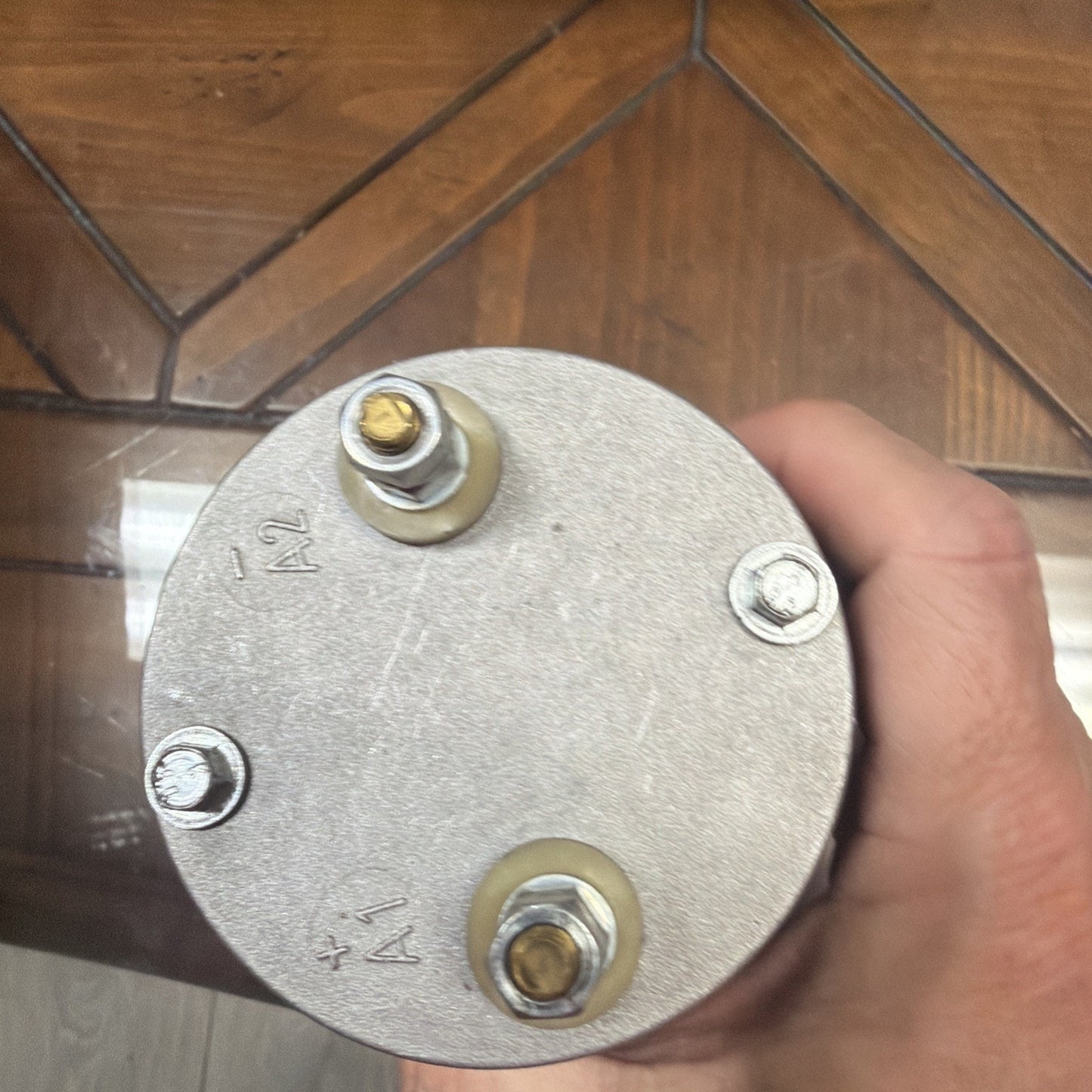 Top view of a circular metallic component with two large brass terminal bolts labeled "A1" and "A2" and two smaller screws, held by a hand over a wooden surface.