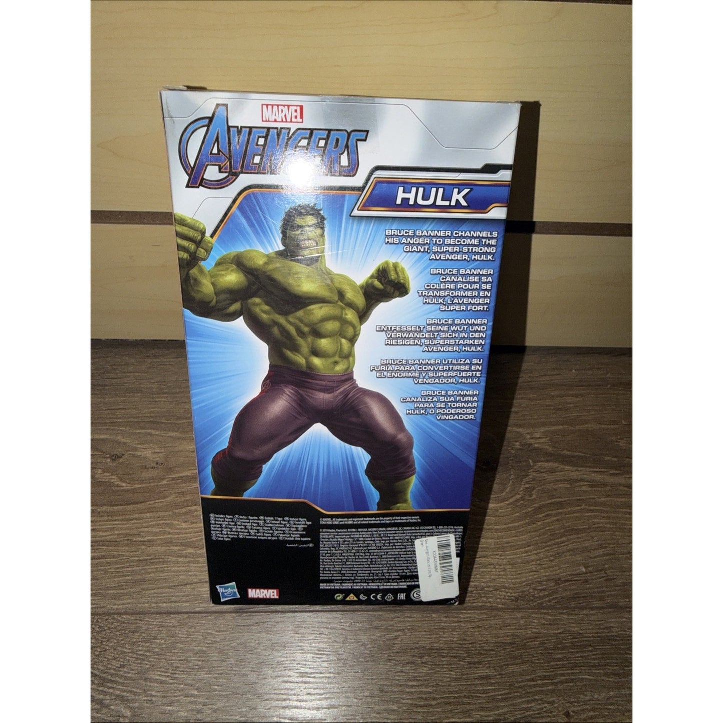 Box back of a Marvel Avengers Hulk action figure featuring an illustrated muscular Hulk in purple pants with text describing the character and product details on a wooden surface.