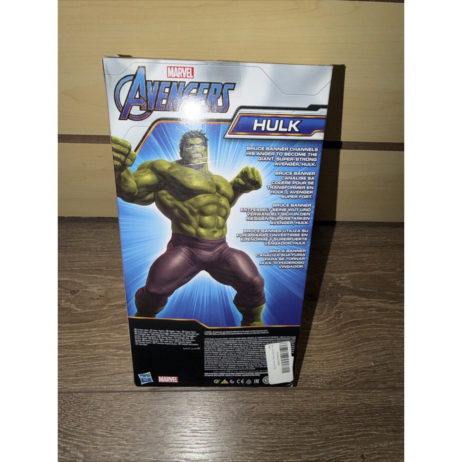 Box back of a Marvel Avengers Hulk action figure featuring an illustrated muscular Hulk in purple pants with text describing the character and product details on a wooden surface.