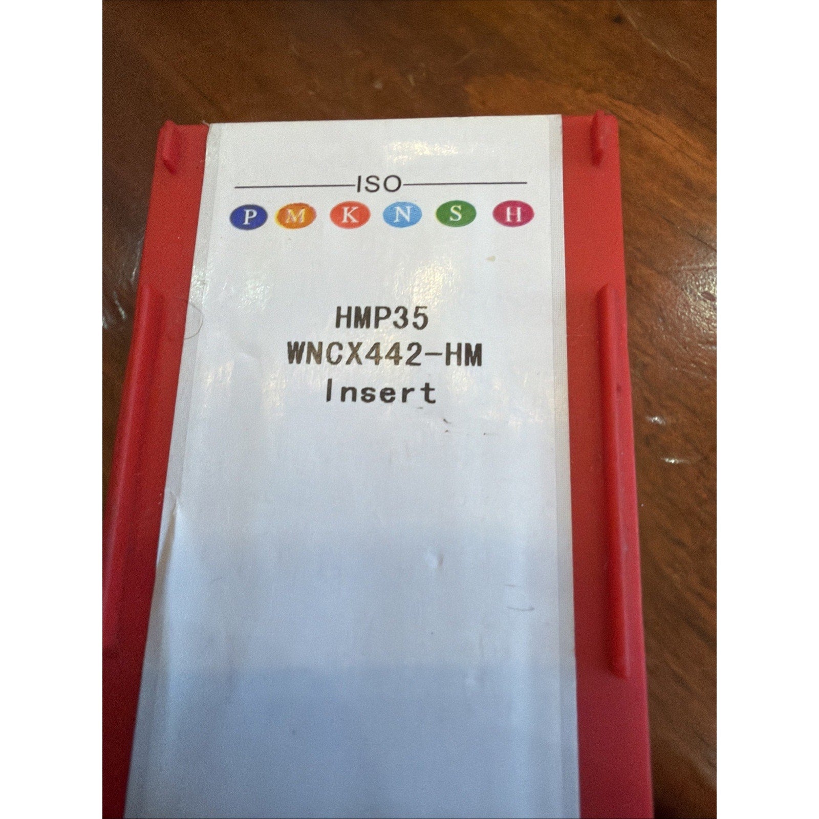 Red plastic container with a white label displaying colored ISO classification symbols and text reading "HMP35 WNCX442-HM Insert" on a wooden surface.