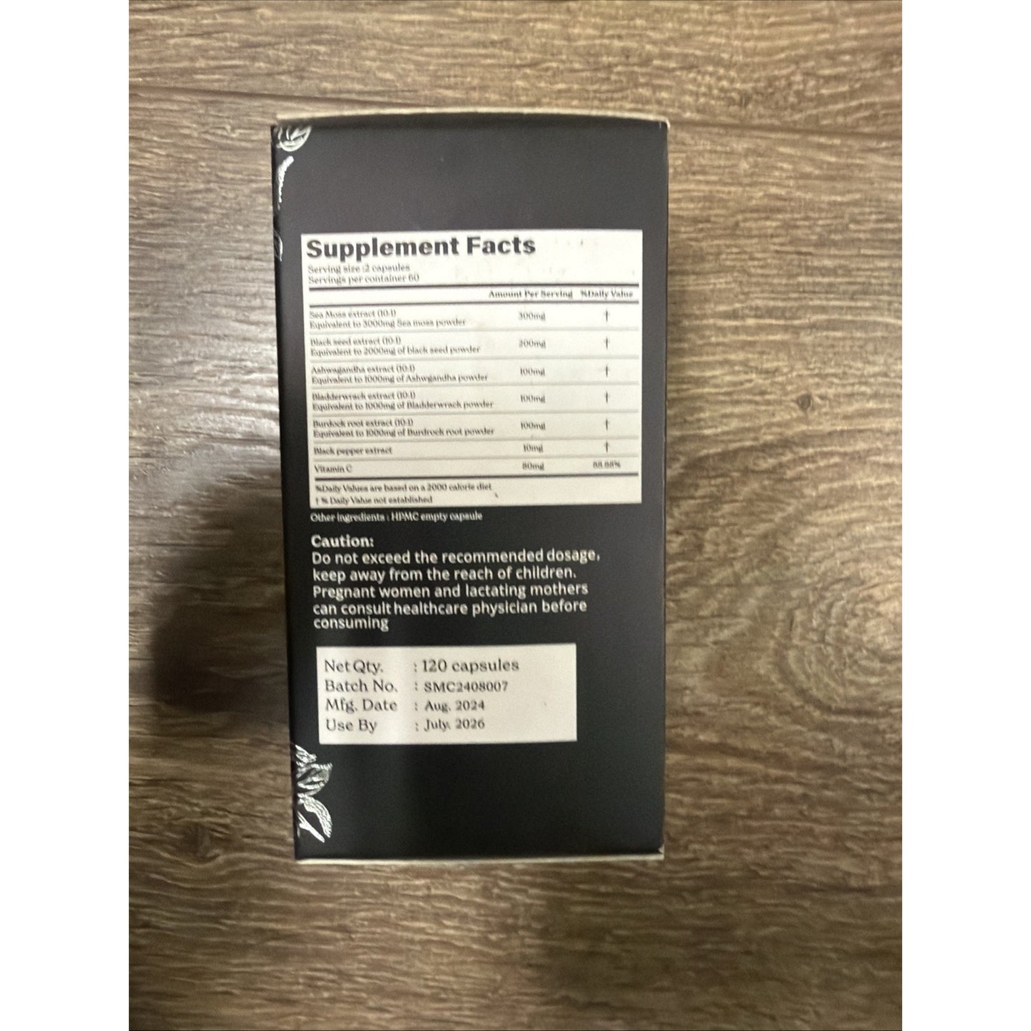 Black supplement bottle label listing nutritional facts including sea moss, black seed, ashwagandha, and black pepper with caution text, batch number, manufacturing and expiration dates on a wooden surface.