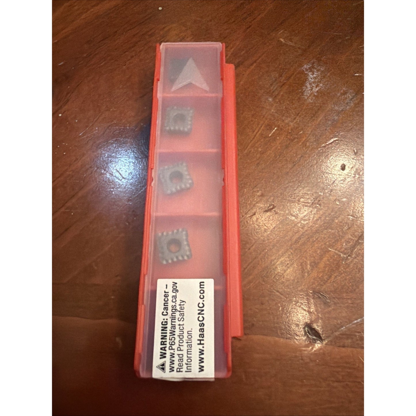 Red rectangular plastic container with a translucent lid holding four square metal cutting inserts with central holes, placed on a wooden surface showing natural grain and a warning label visible inside the lid.