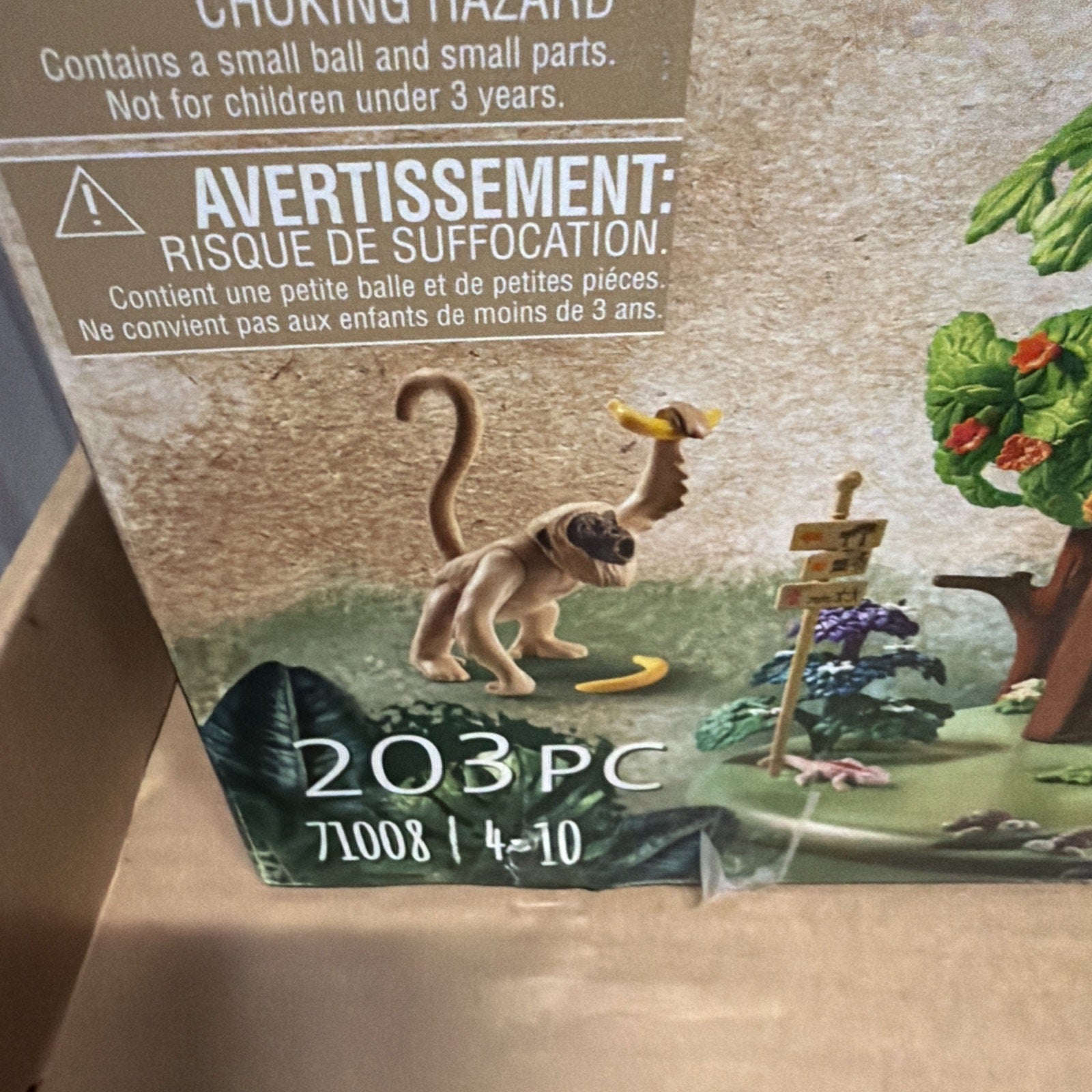 Close-up of a toy packaging corner showing a small monkey figure holding a banana, a wooden signpost, and part of a tree with green foliage, with text warning about choking hazards and indicating 203 pieces.
