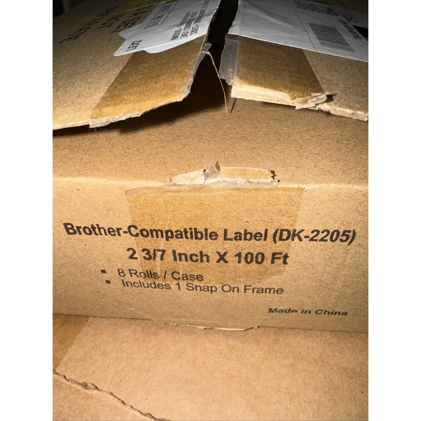 Brown corrugated cardboard box with printed text indicating Brother-compatible label rolls (DK-2205), dimensions 2 3/7 inch by 100 feet, including 8 rolls per case and 1 snap-on frame, with visible shipping labels and partially opened flaps.