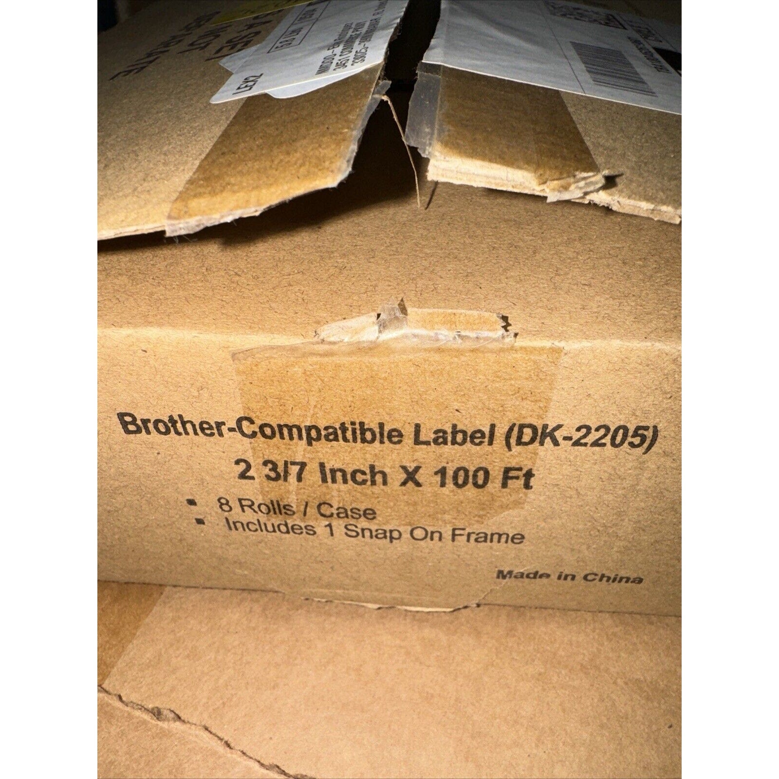 Brown corrugated cardboard box with printed text indicating Brother-compatible label rolls (DK-2205), dimensions 2 3/7 inch by 100 feet, including 8 rolls per case and 1 snap-on frame, with visible shipping labels and partially opened flaps.