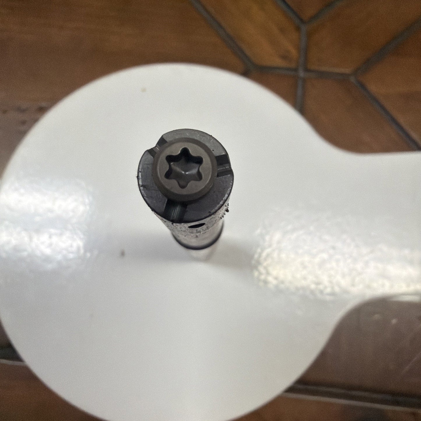 Top-down view of a black metal tool with a star-shaped tip resting on a glossy white circular surface, with a brown tiled floor in the background.