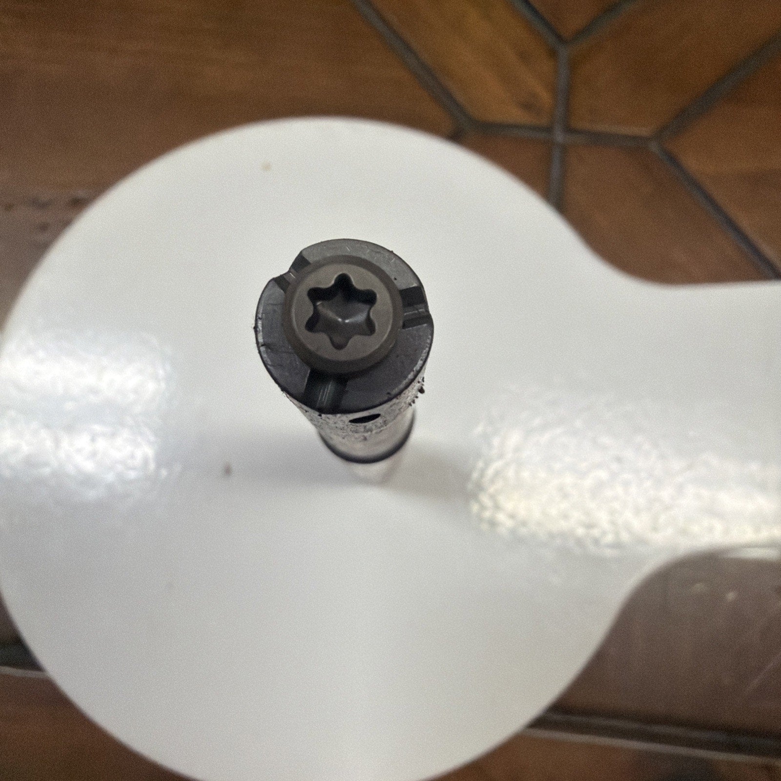 Top-down view of a black metal tool with a star-shaped tip resting on a glossy white circular surface, with a brown tiled floor in the background.