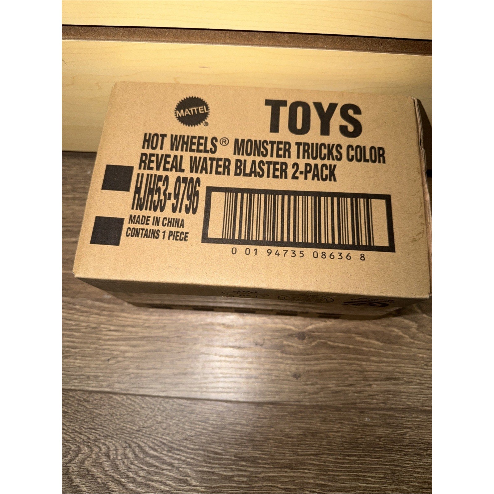 Brown cardboard box labeled with black text indicating a Hot Wheels Monster Trucks Color Reveal Water Blaster 2-pack, featuring a barcode and made in China details on a wooden floor.