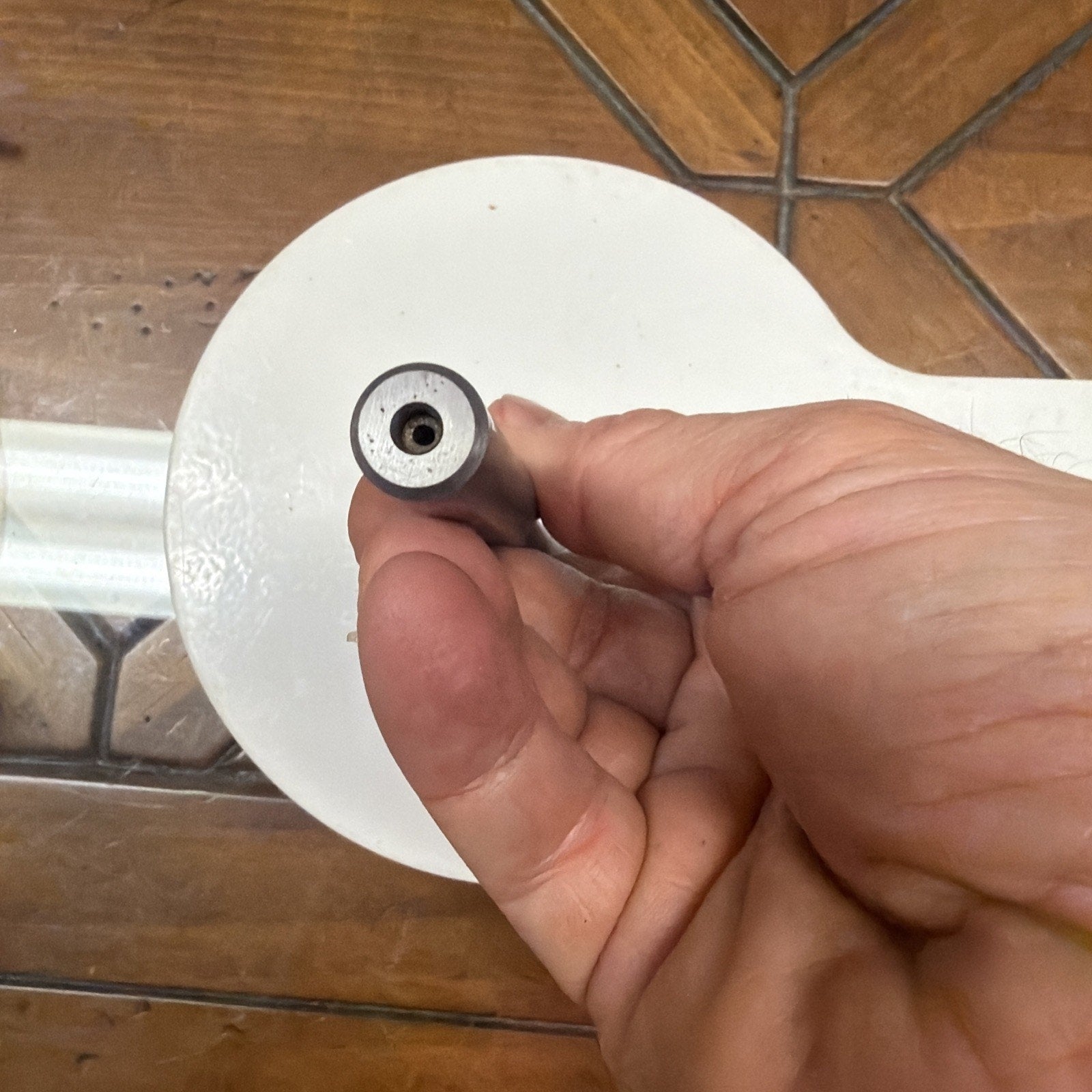 Close-up of a hand holding a small cylindrical metal object with a hollow center, positioned over a white circular surface and a brown tiled floor background.