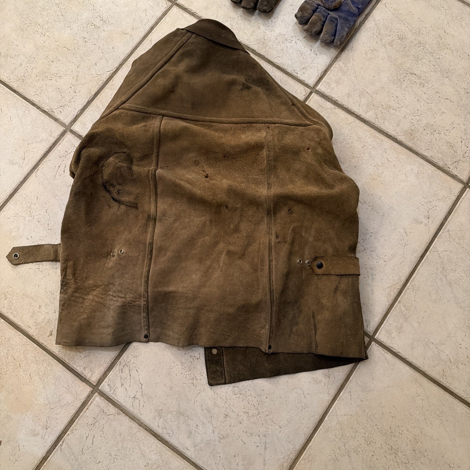 Heavy-duty brown leather welding jacket coat with reinforced seams and adjustable strap laid flat on tiled floor