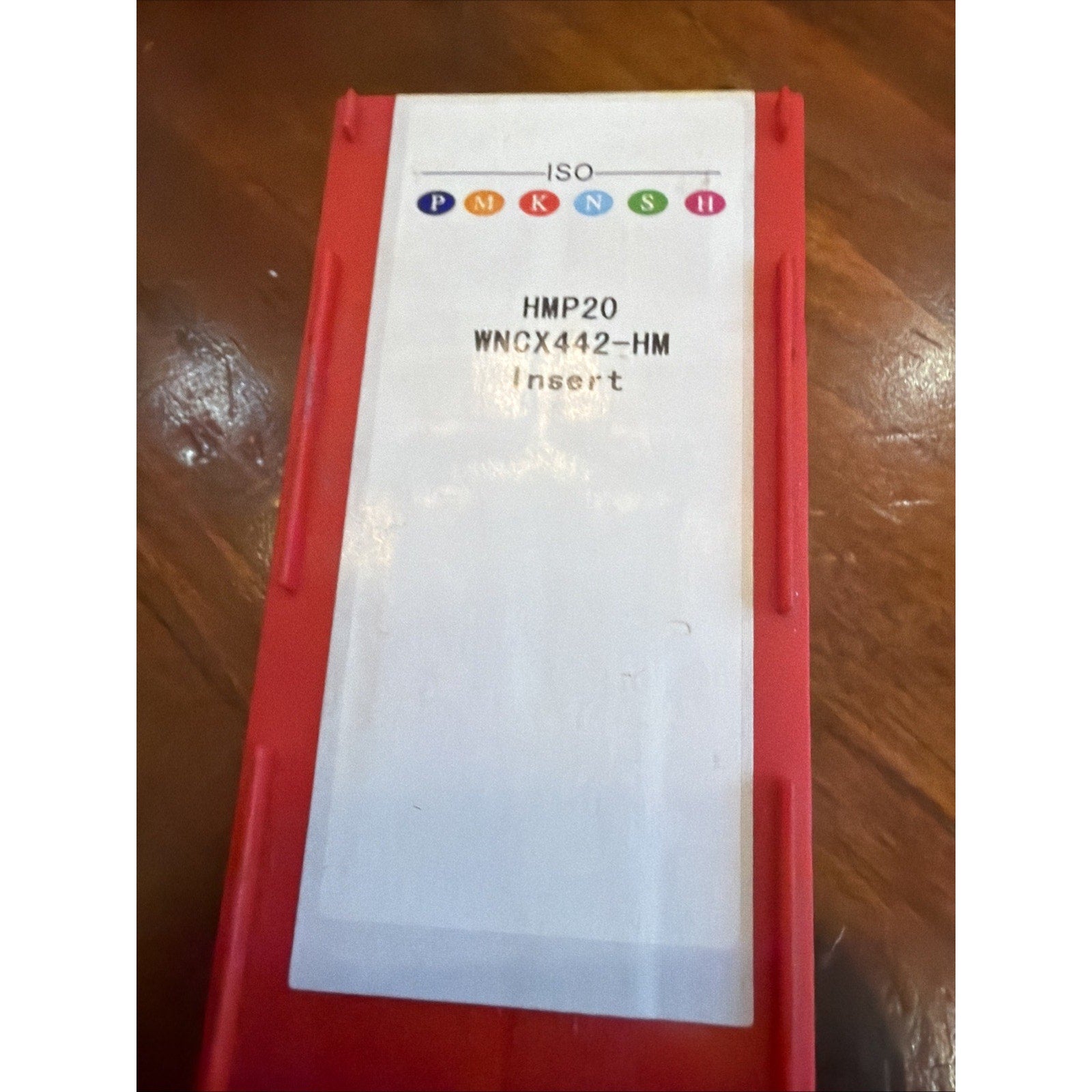 Rectangular red plastic container with a white label displaying colored ISO standard symbols and text reading "HMP20 WNCX442-HM Insert" on a wooden surface.