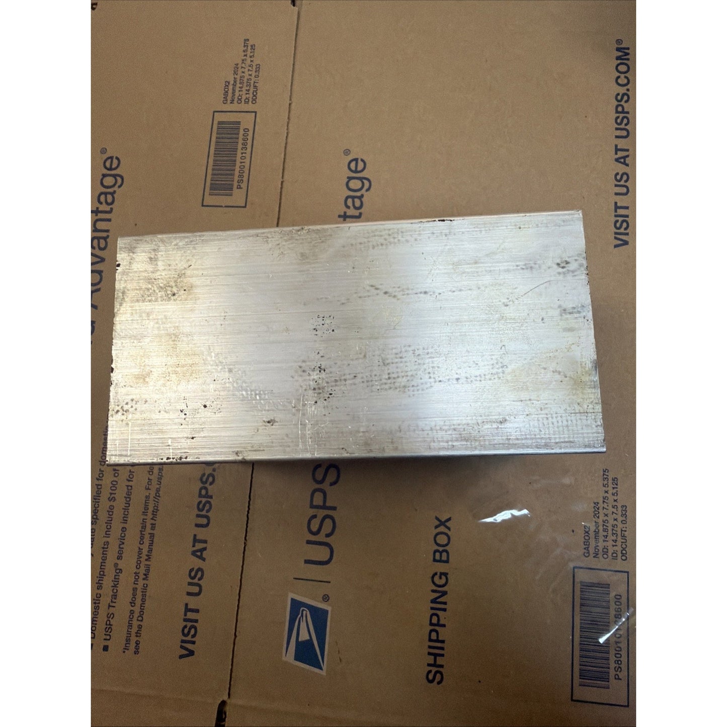 Rectangular piece of metal with a brushed surface finish, showing scratches and discoloration, resting on a cardboard shipping box with USPS branding.