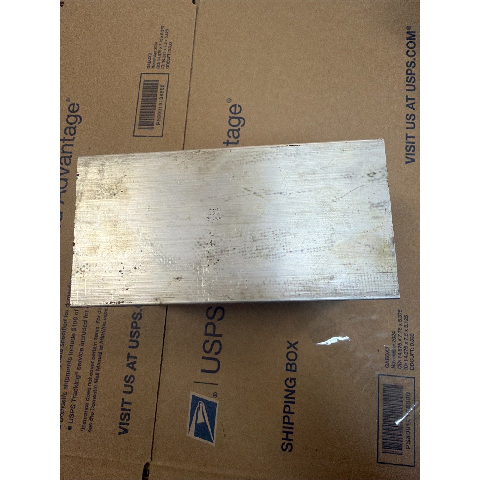 Rectangular piece of metal with a brushed surface finish, showing scratches and discoloration, resting on a cardboard shipping box with USPS branding.