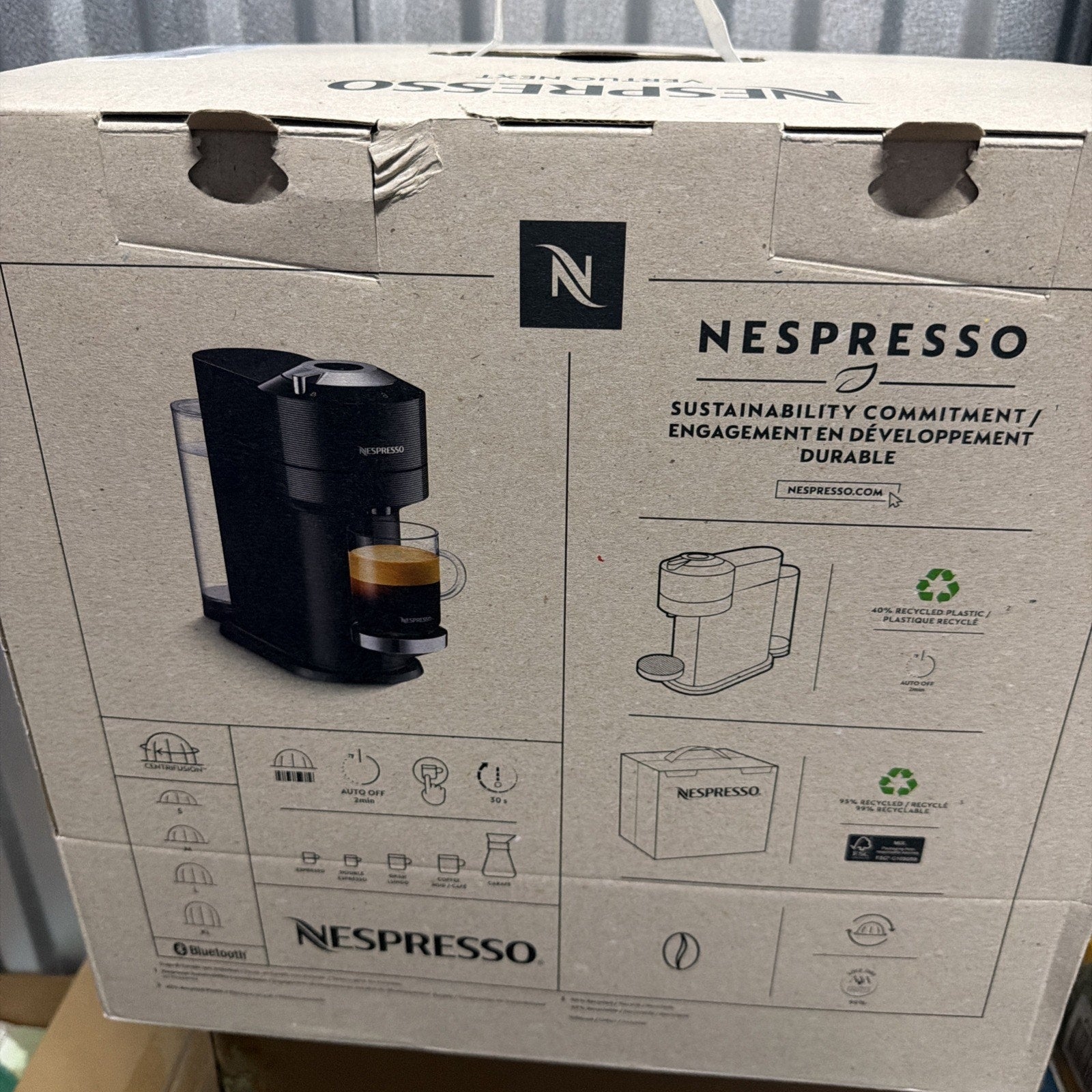 Cardboard packaging for a Nespresso coffee machine featuring a photo of the black machine with a cup of espresso, icons for device features, and text highlighting sustainability and recyclable materials.