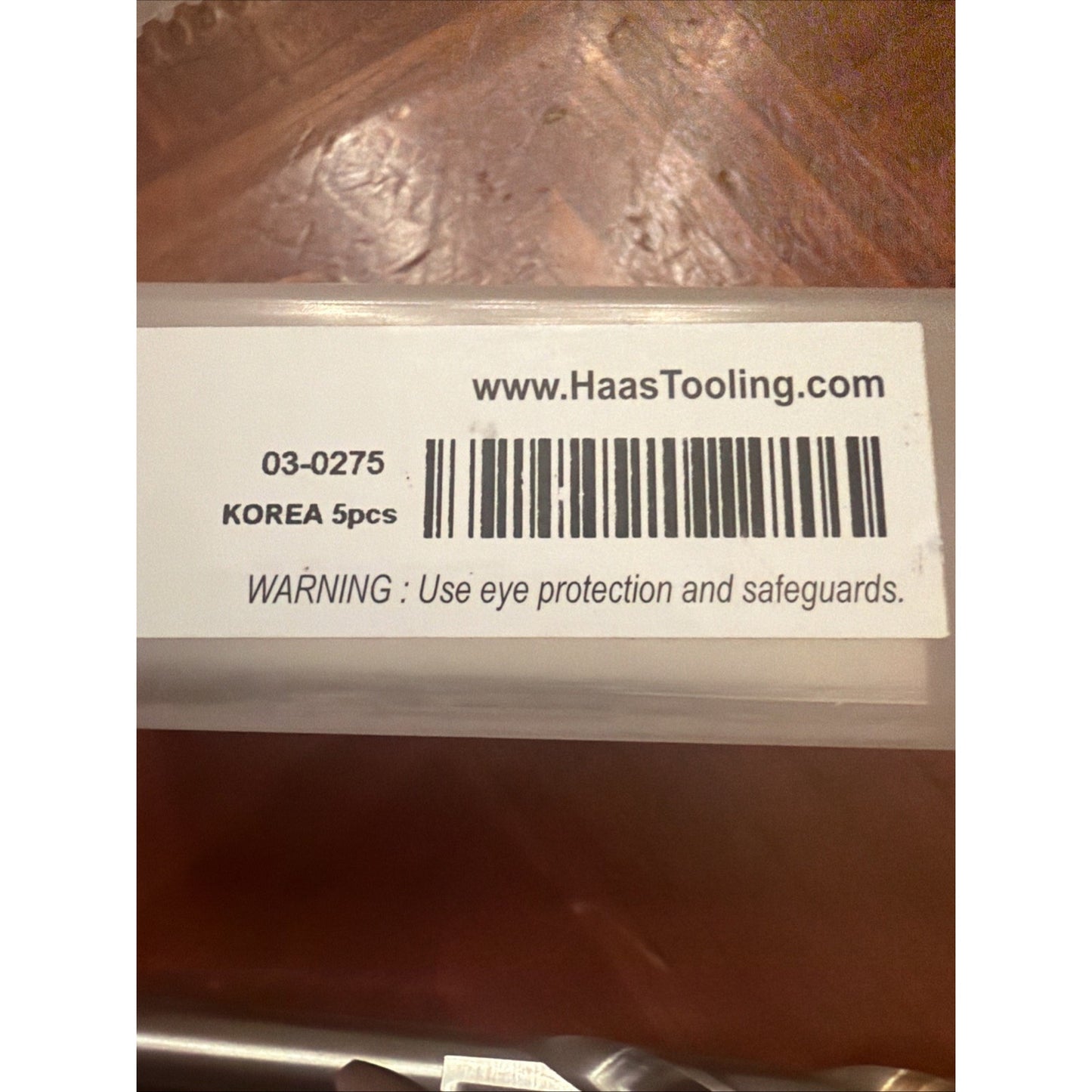 Close-up of a white label on a clear plastic container displaying a barcode, product code 03-0275, origin Korea, quantity 5 pieces, and a safety warning to use eye protection and safeguards.