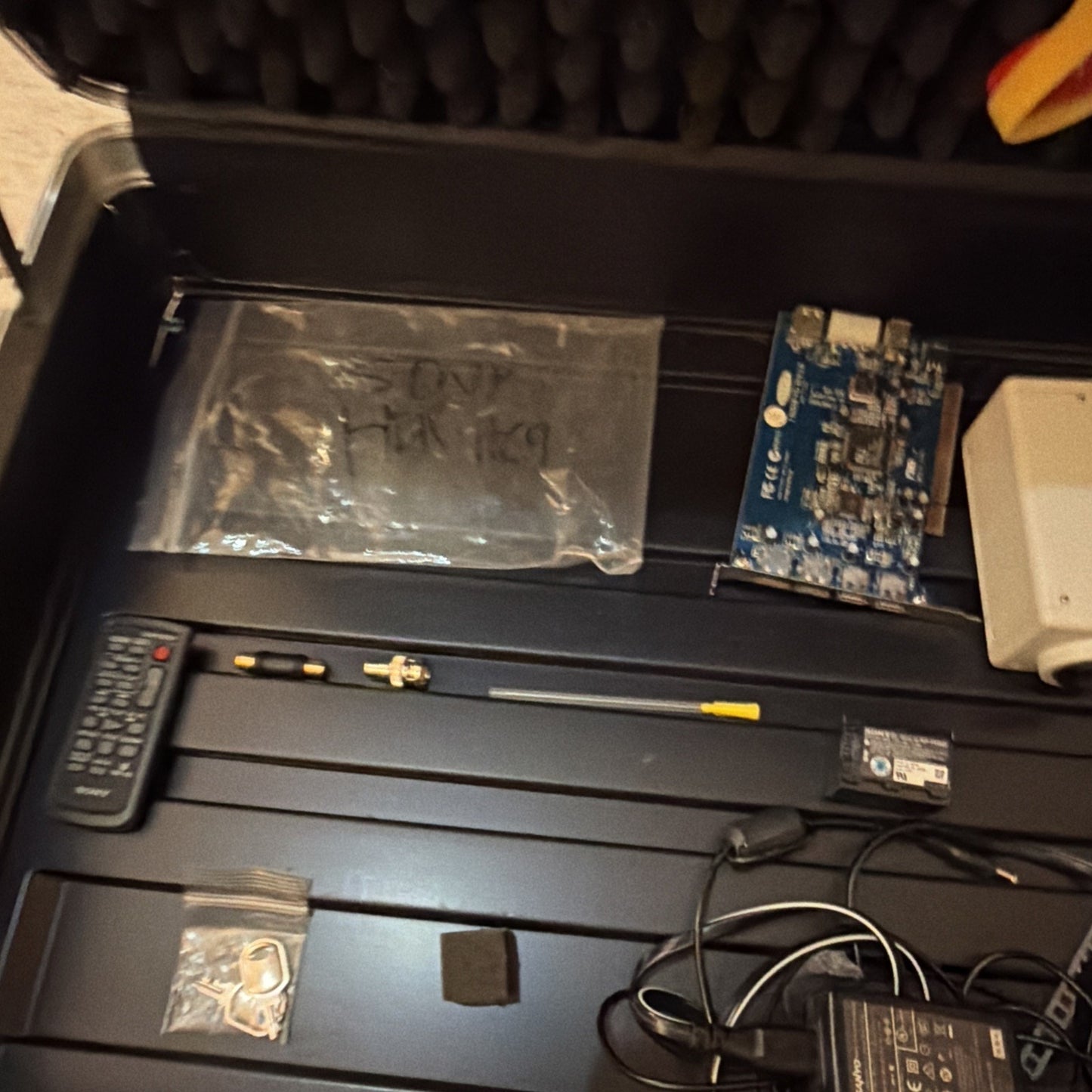Black foam-lined equipment case containing a small black remote control, a long antenna with connectors, a blue circuit board, a white plastic device, various cables, and small plastic bags with metal keys and parts.