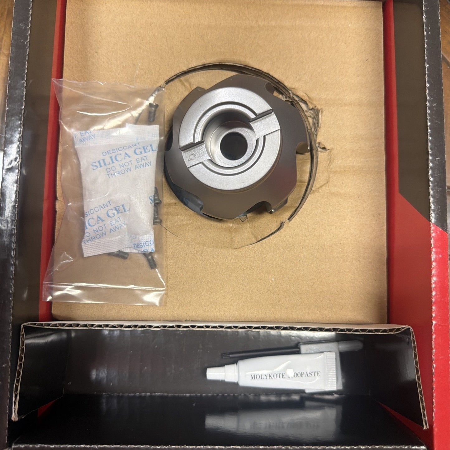 Metal circular tool component with a central hole, packaged in a cardboard box alongside silica gel packets and a small white tube labeled "MOLYKOTE 1000 PASTE" on a black cardboard insert.