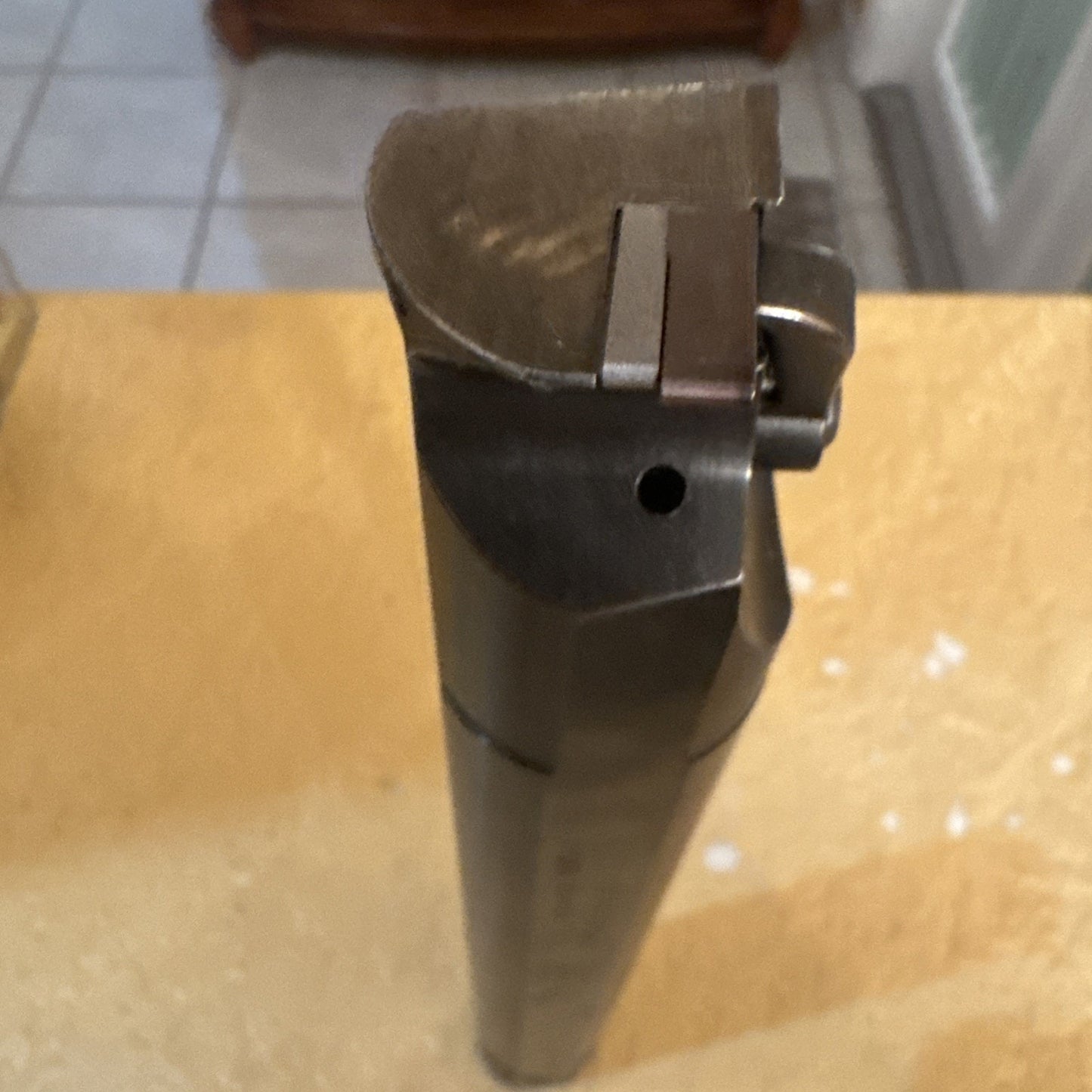 Top-down view of a dark metal cutting tool with a curved cutting edge and a small hole on the side, placed vertically on a yellowish surface with a tiled floor in the background.