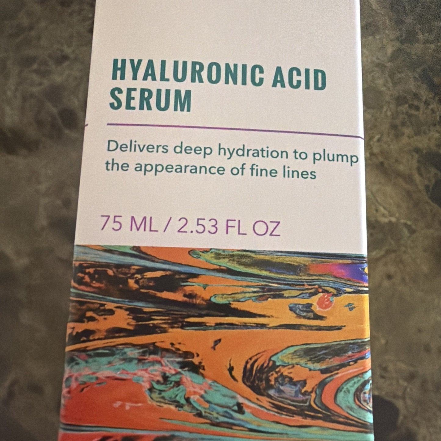 Close-up of skincare serum packaging featuring bold teal text stating "Hyaluronic Acid Serum" with a description highlighting deep hydration to reduce fine lines, and a colorful abstract marbled design along the bottom edge.
