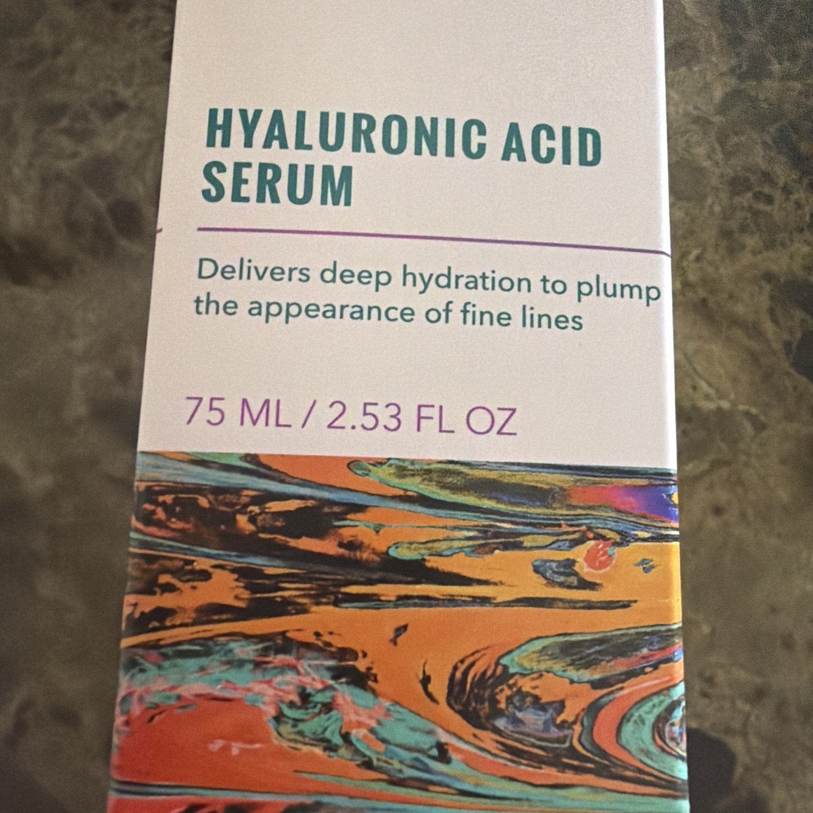 Close-up of skincare serum packaging featuring bold teal text stating "Hyaluronic Acid Serum" with a description highlighting deep hydration to reduce fine lines, and a colorful abstract marbled design along the bottom edge.
