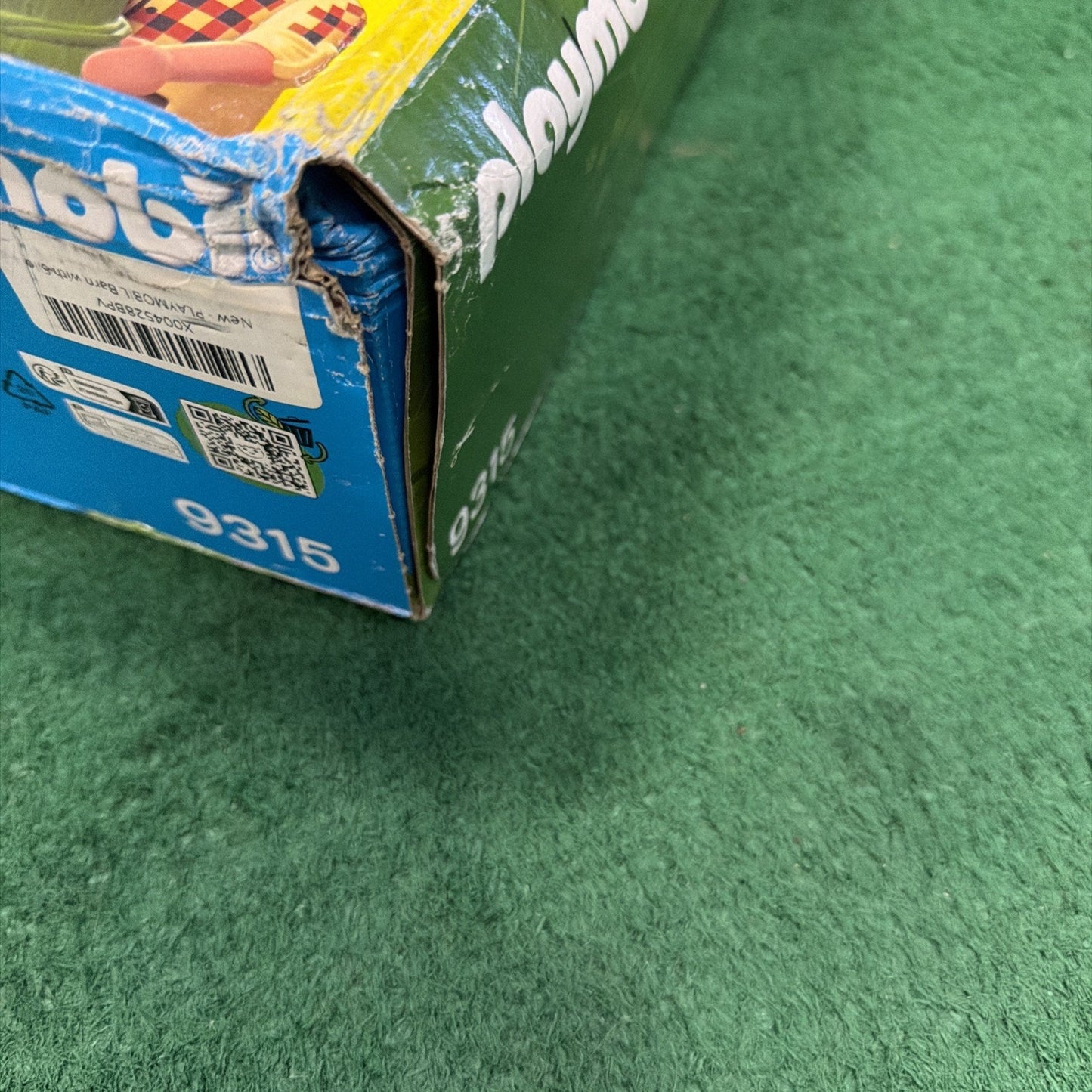 Corner of a worn Playmobil toy box with visible creases and tears, resting on a green textured surface.