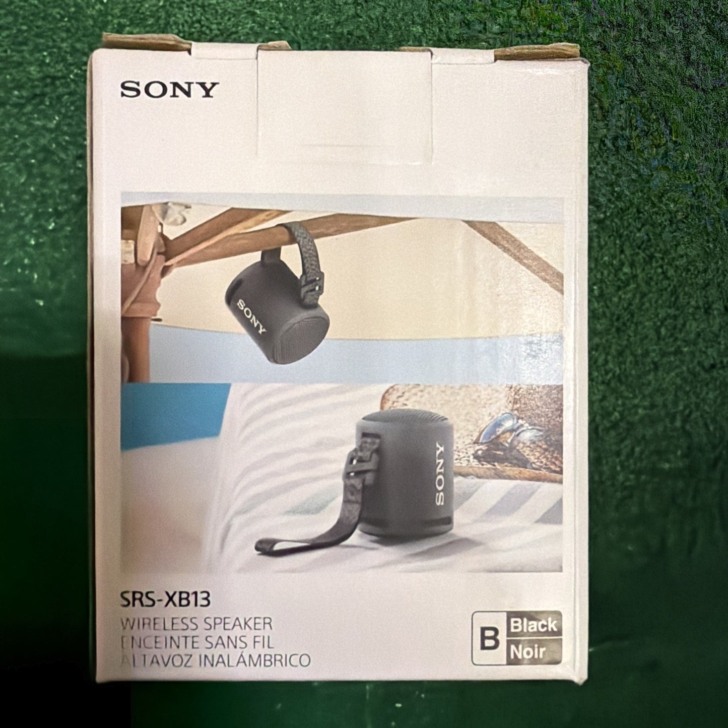 Sony branded white packaging box featuring images of a compact black wireless speaker with a carrying strap hanging from a wooden beam and placed on a striped surface indoors.