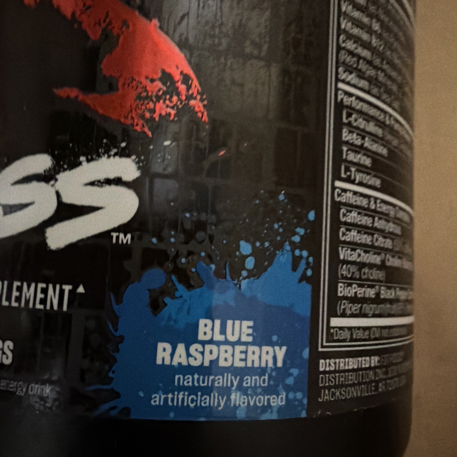 Close-up of a black supplement container label highlighting blue raspberry flavor with natural and artificial ingredients, featuring blue paint splatter graphic and partial nutritional facts.