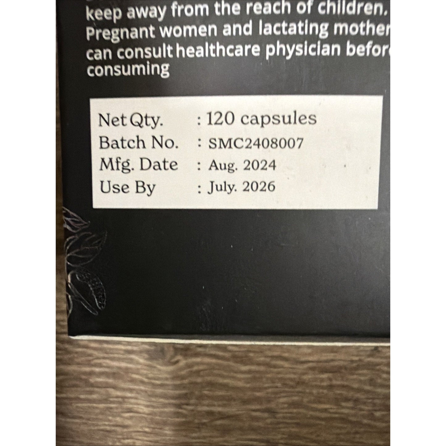Close-up of a black product box label showing batch number SMC2408007, manufacturing date August 2024, use-by date July 2026, and quantity of 120 capsules on a wooden surface.