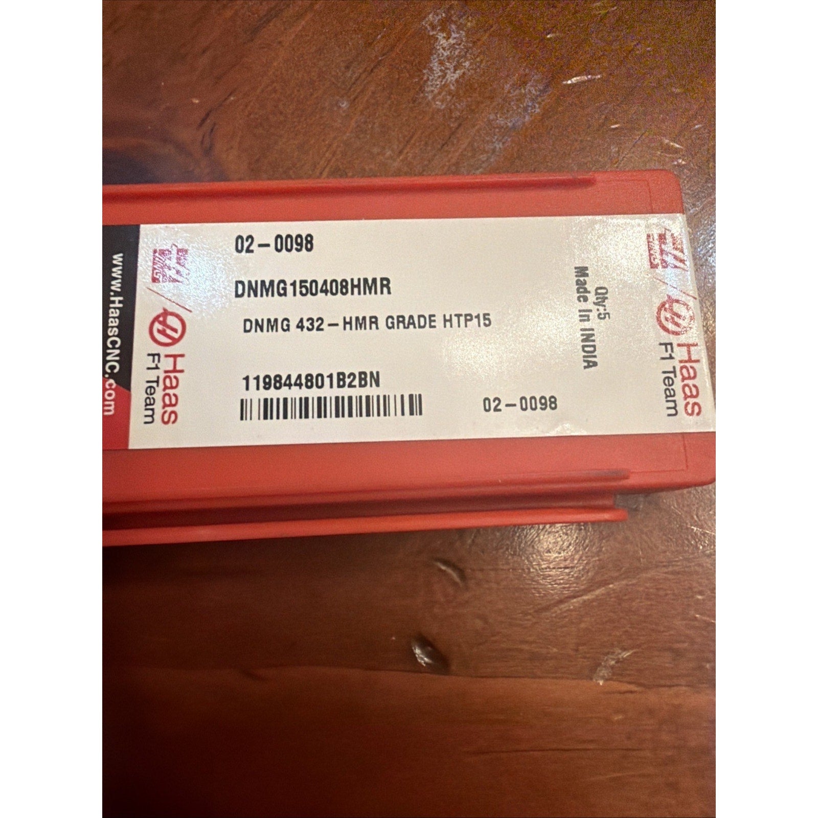 Red plastic rectangular container with a white label showing product code "DNMG150408HMR," grade "HTP15," barcode, and "Made in India," placed on a scratched wooden surface.