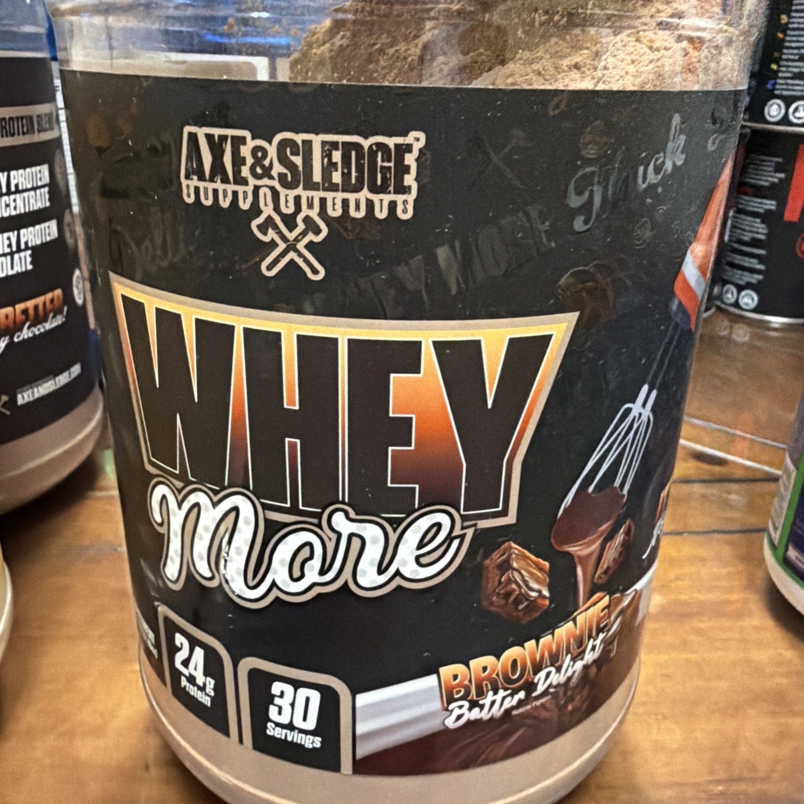 Large black container of Axe & Sledge Whey More protein powder in brownie butter dulce flavor, featuring 24 grams of protein and 30 servings per container, with a chocolate drizzle and dessert pieces illustration.