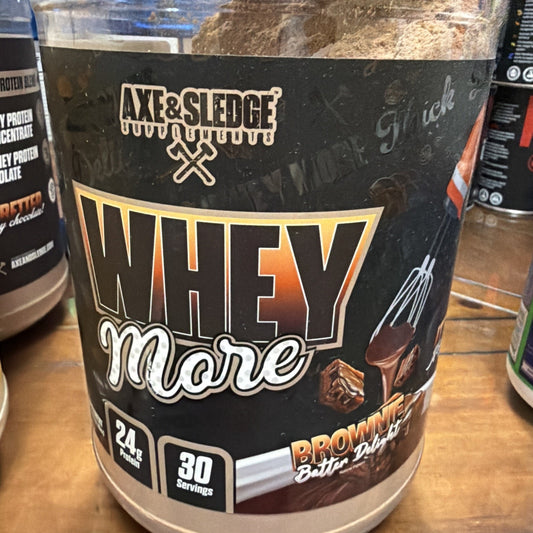 Large black container of Axe & Sledge Whey More protein powder in brownie butter dulce flavor, featuring 24 grams of protein and 30 servings per container, with a chocolate drizzle and dessert pieces illustration.
