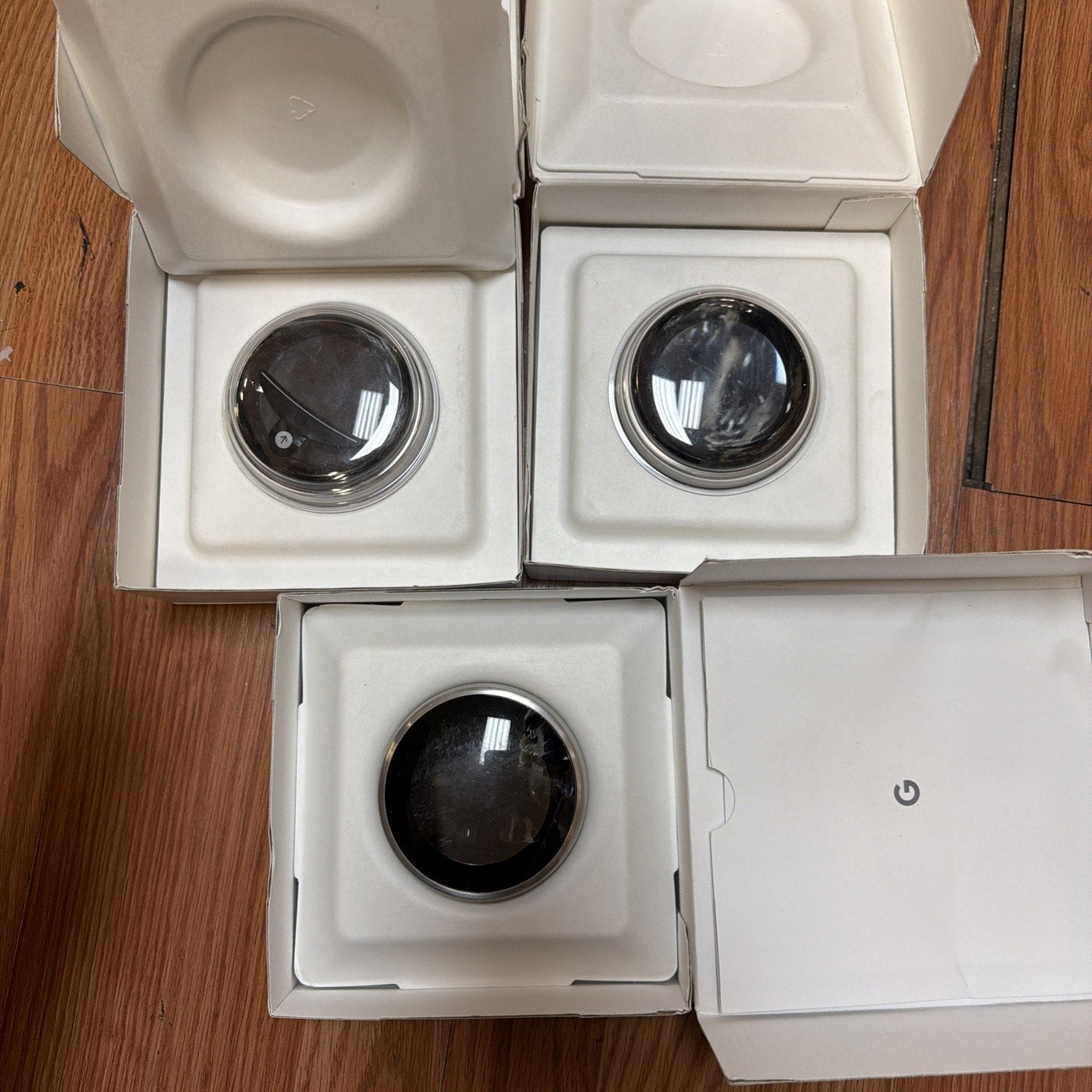 Three smart thermostat devices with round black screens are displayed inside white square protective boxes on a wooden surface.