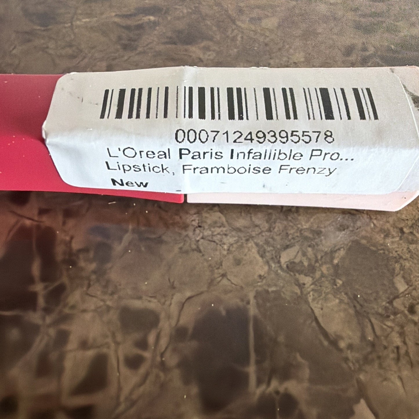 Close-up of a white label with black barcode and text showing "L'Oreal Paris Infallible Pro Lipstick, Framboise Frenzy, New" attached to a pink lipstick tube on a brown textured surface.