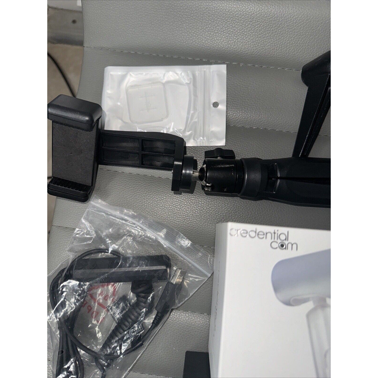 Lot of camera accessories including phone mount tripod, cables in plastic bag, and Credential Cam box on gray surface