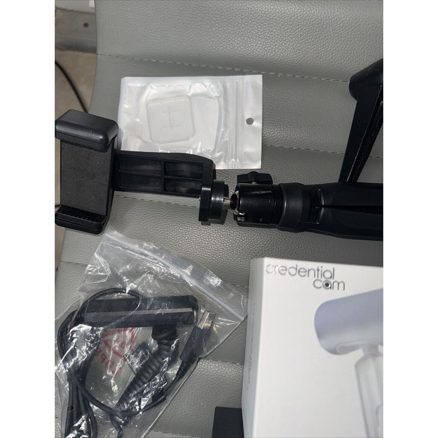 Lot of camera accessories including phone mount tripod, cables in plastic bag, and Credential Cam box on gray surface