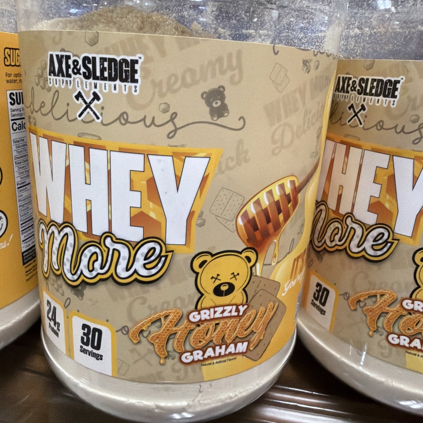 Clear plastic container of Axe & Sledge Whey More protein powder in Grizzly Honey Graham flavor, with label highlighting 24 grams of protein and 30 servings per container, featuring a honey dipper and teddy bear illustration.